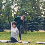 Diligent yoga student is learning Cow Face position sitting on mat and stretching arms while experienced teacher is helping her, girls are talking and laughing at outdoor ptactice.
